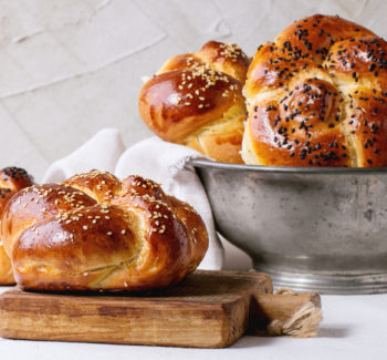 Heap of sweet round sabbath challah bread with white and black sesame seeds in vintage metal bowl and on small cutting board over white table with plastered wall at background.